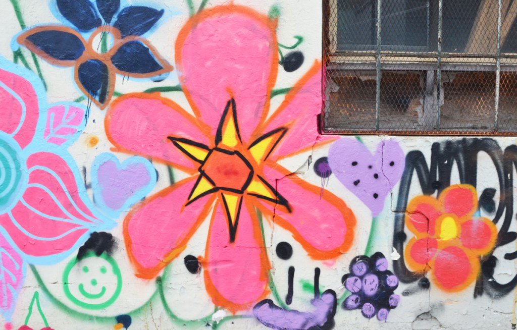 painted, large 6 petal pink flower with yellow triangles at central edge, and an orange hexagon center, on same wall, a green happy face 