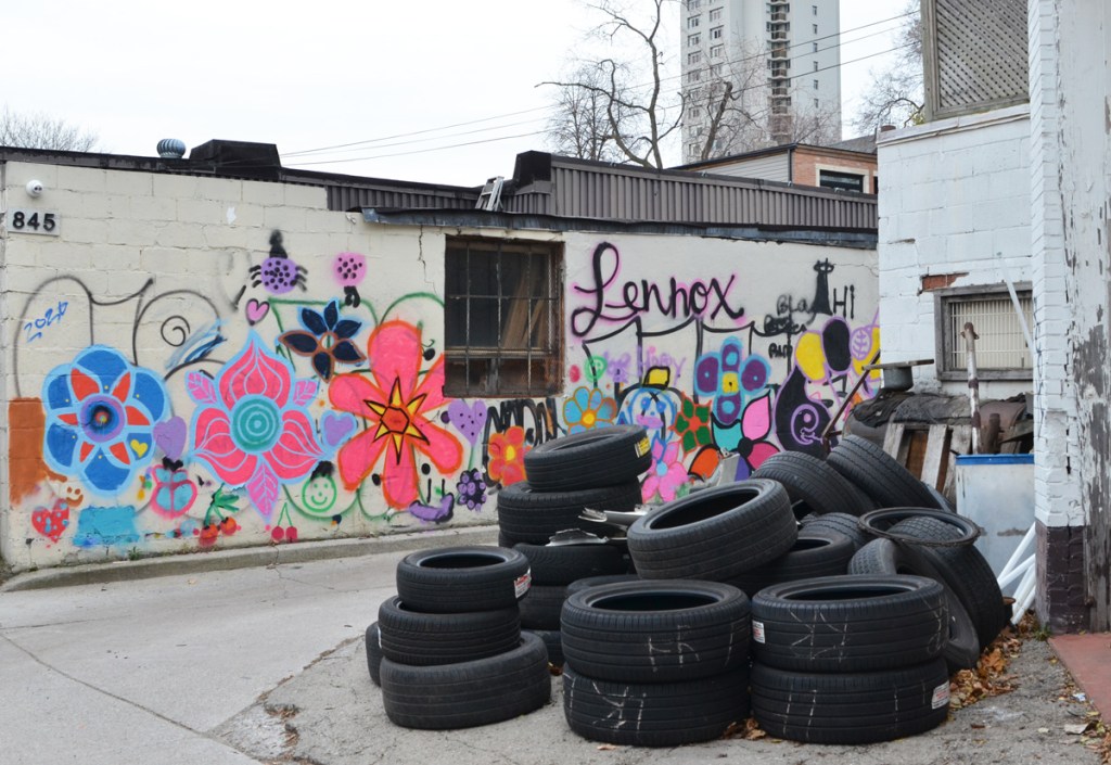 a pile of tires in an alley across from a white wall painted with lots of colourful flowers, stylized form 