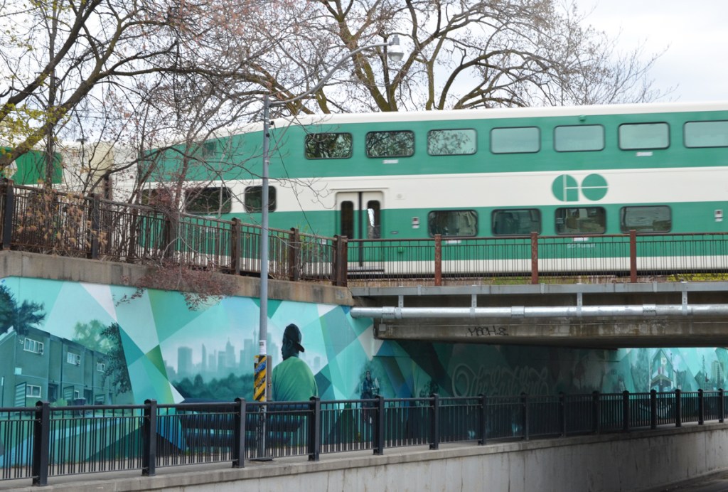 green and white GO train passing over bridge over Jones Ave. where a mural in shades of green and blue has been painted 