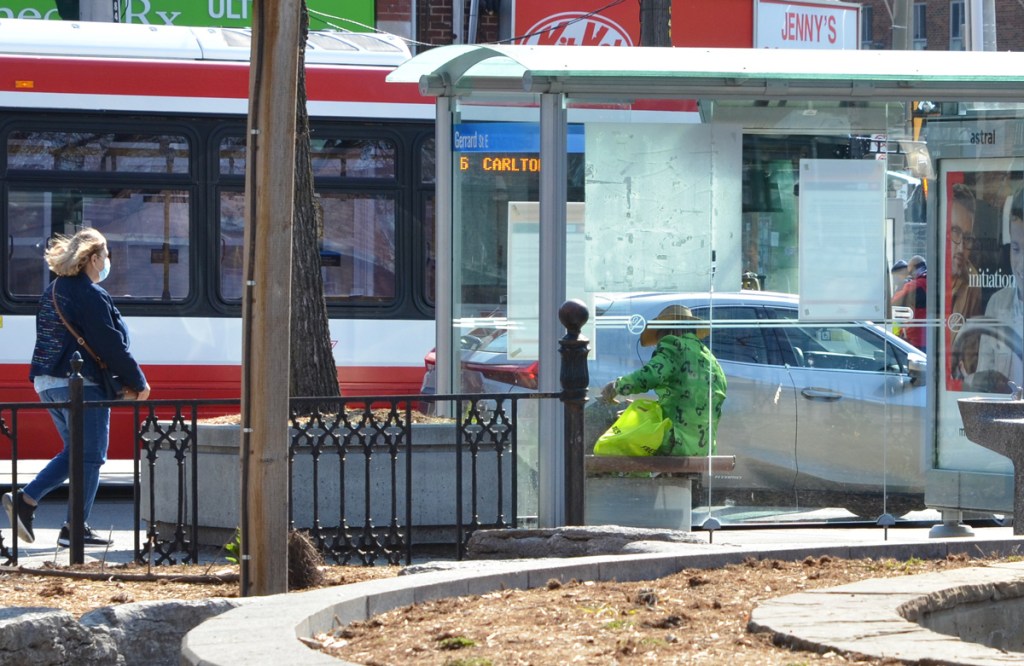 Man in a green jacket with dark green question marks all over it like the joker from Batman sits in a bus shelter as a woman walks past