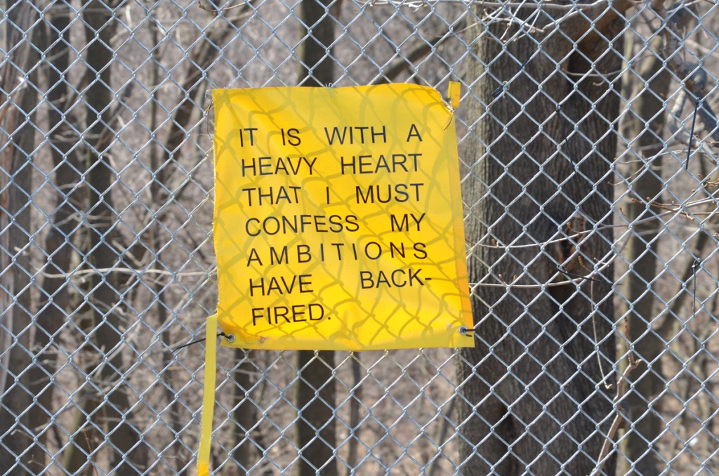 yellow poster with words on a chain link fence  words say it is with a heavy heart that i must confess my ambitions have backfired