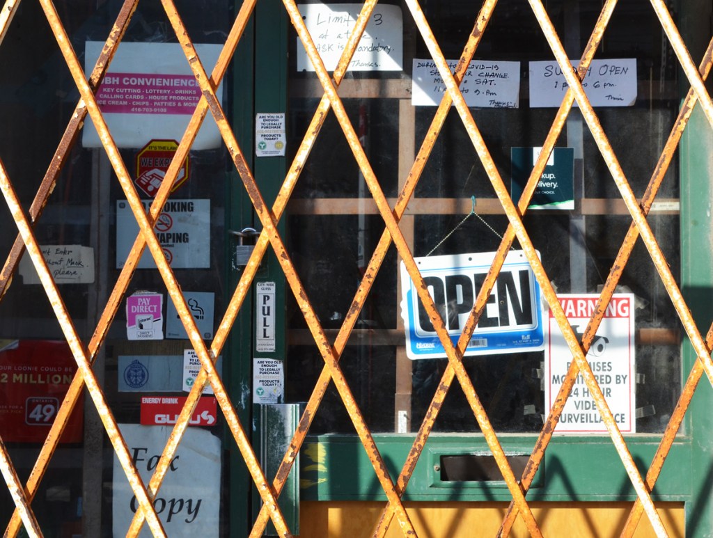 rusty yellow metal grille closed in front of a store front with an open sign, ans some had written signs re opening hours