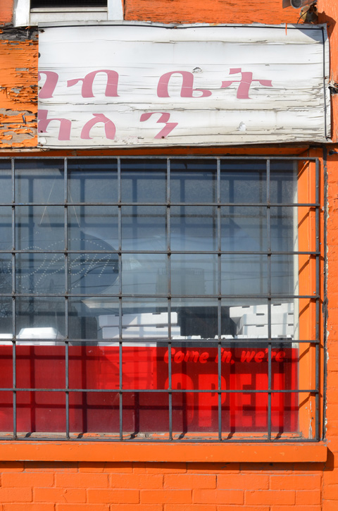 orange storefront with window.  metal grille over window, red plastic in bottom of window, broke sign in different alphabet over the window 