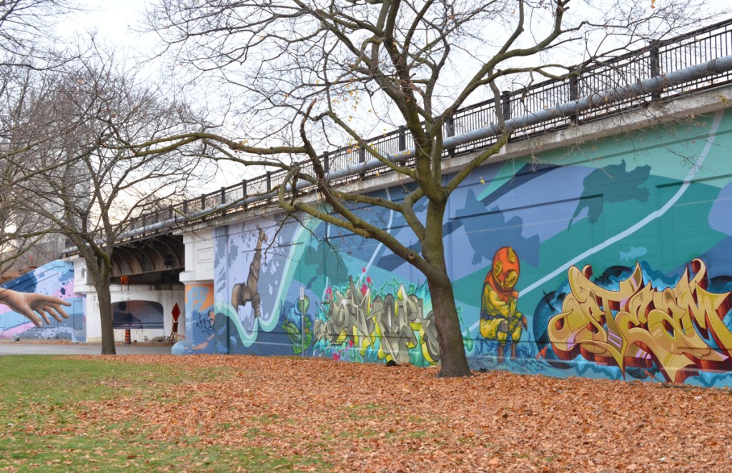mural on a wall along railway embankment, park in front of wall.  water theme mural, large faucet has water running out of it, a frog, a scuba diver, and some text street art 