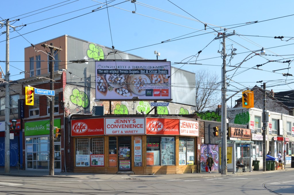 northeast corner of parliament and gerrard with Jennys convenience store with its red and white sign with kit kat advertising, billboard above the store, other stores adjacent to it, Jerk restaurant, Remedy Rx, 
