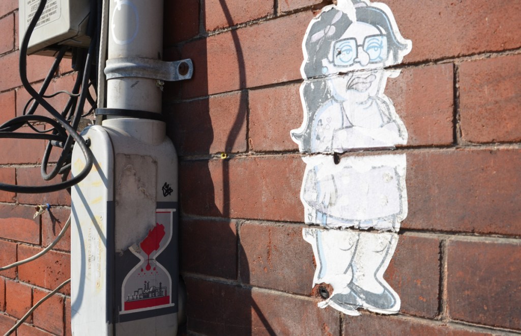 A small paste up on a brick wall, black and white drawing of a woman in black flats, shorts, sleeveless top, square glasses, and long black hair. There is also a lovebot sticker nearby, an egg timer with red heart on top and city of Toronto skyline on the bottom