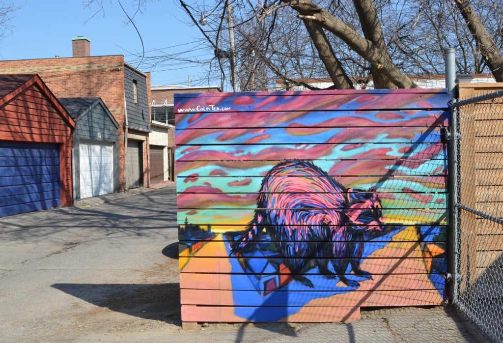 a  mural of a larger than life raccoon standing over houses, by Colin Tea, in a lane with other garages