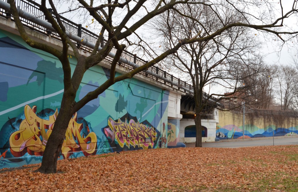 park in autumn, wall of railway embankment runs along edge of park, mural on the wall, water theme, with text throw up street art by tenser and steam