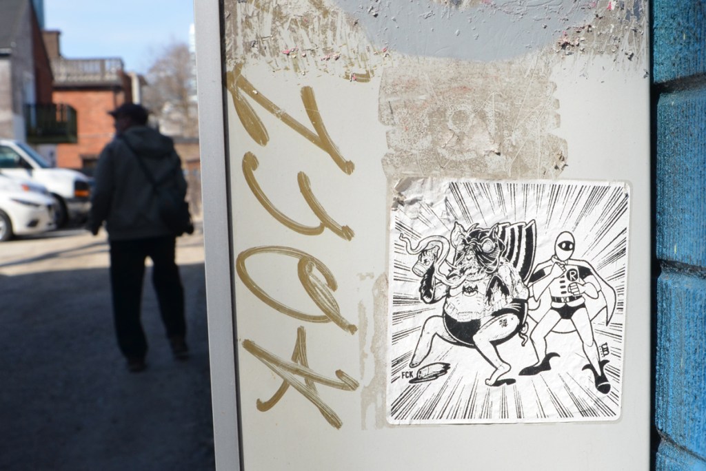 a man walks past a black and white urban ninja squadron sticker,  T bonez dressed as robin and another character dresses as Batman 