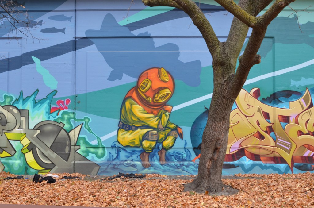 tree in front of a mural, painting of a person in vintage scuba outfit with old fashioned helmet, yellow suit, weight belt, sitting on a box or a rock at the bottom of the lake, silhouette fish swimming past 