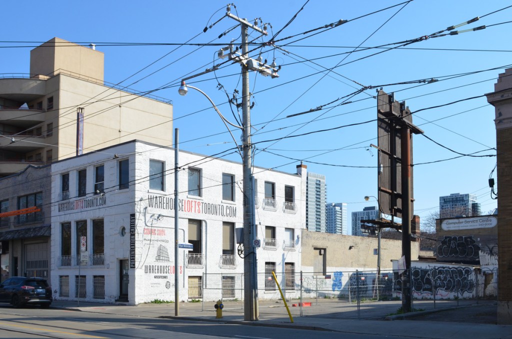 utility pole with many wires, an old brick warehouse that has been converted into loft apartments, a vacant lot surround by fence, a tall billboard seen from the back 