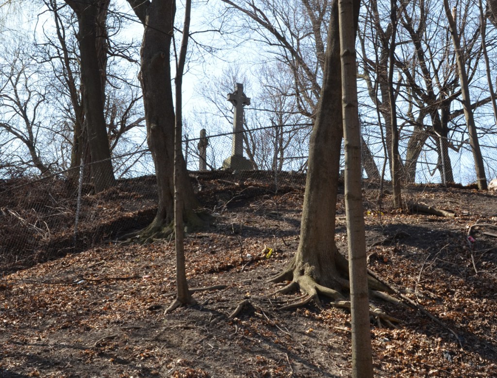 looking uphill through trees to cemetery markers in the cemetery on the other side of a chainlink fence