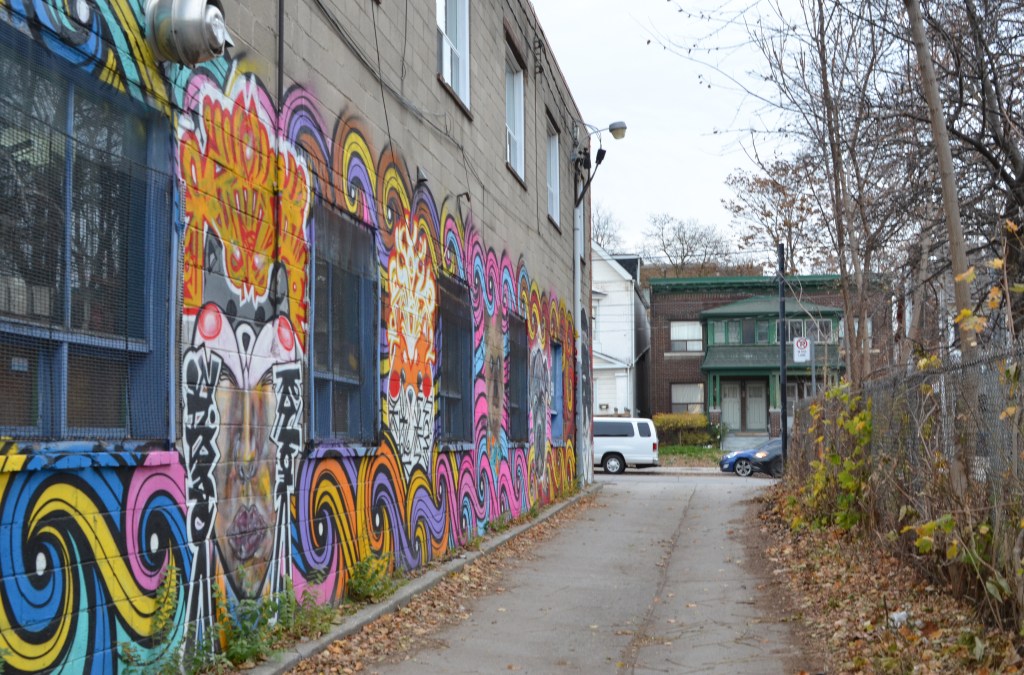 looking up an alley with a mural on the left and a chainlink fence with lots of saplings on the right 