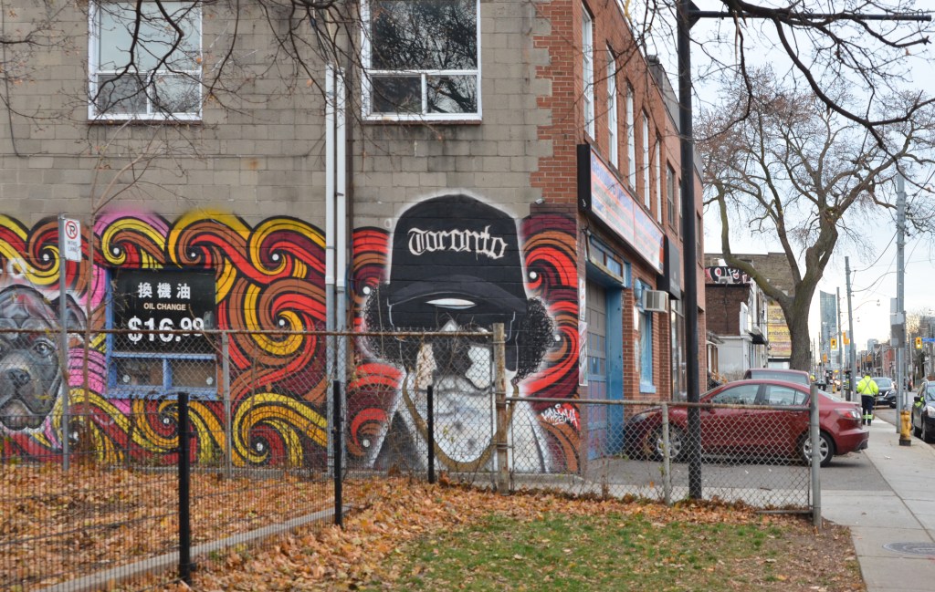 red car parked in front of a body shop with a mural on the side of its building.  Black and white dog in mural with black fuzzy ears, sunglasses and a Toronto baseball cap 