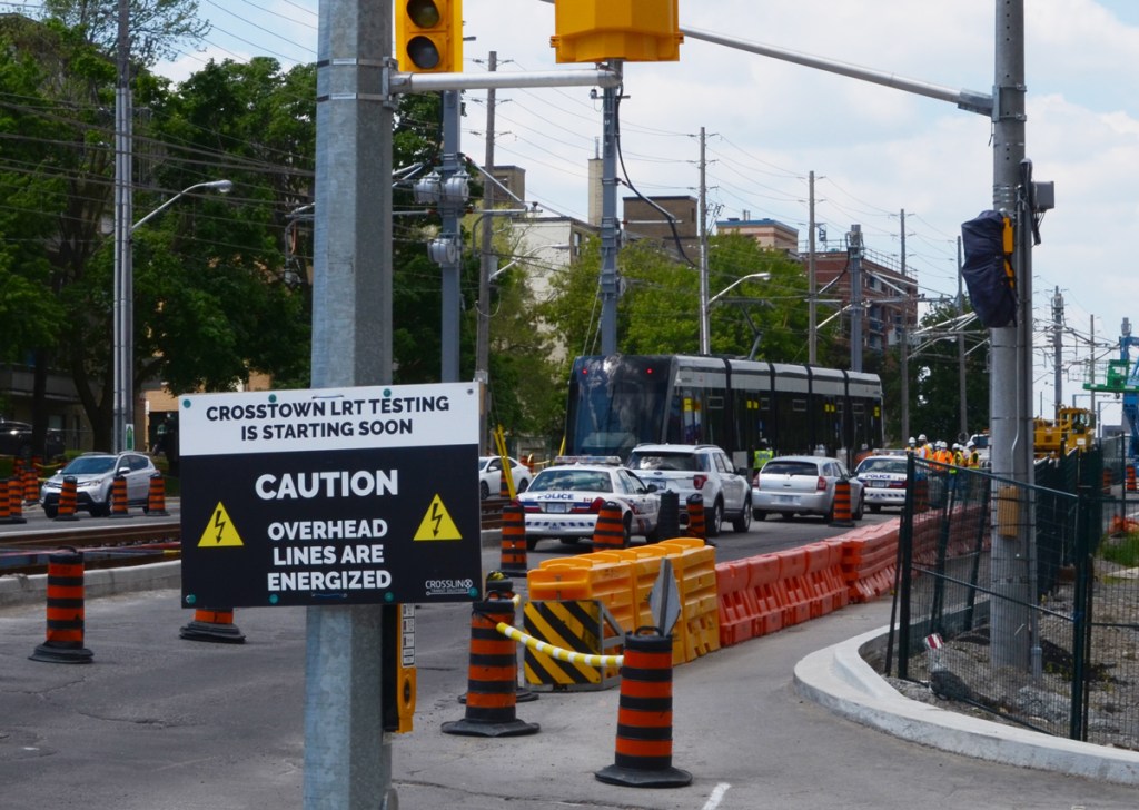 sign saying crosstown LRT testing is starting soon, caution, overhead lines are energized, on street near near new LRT train