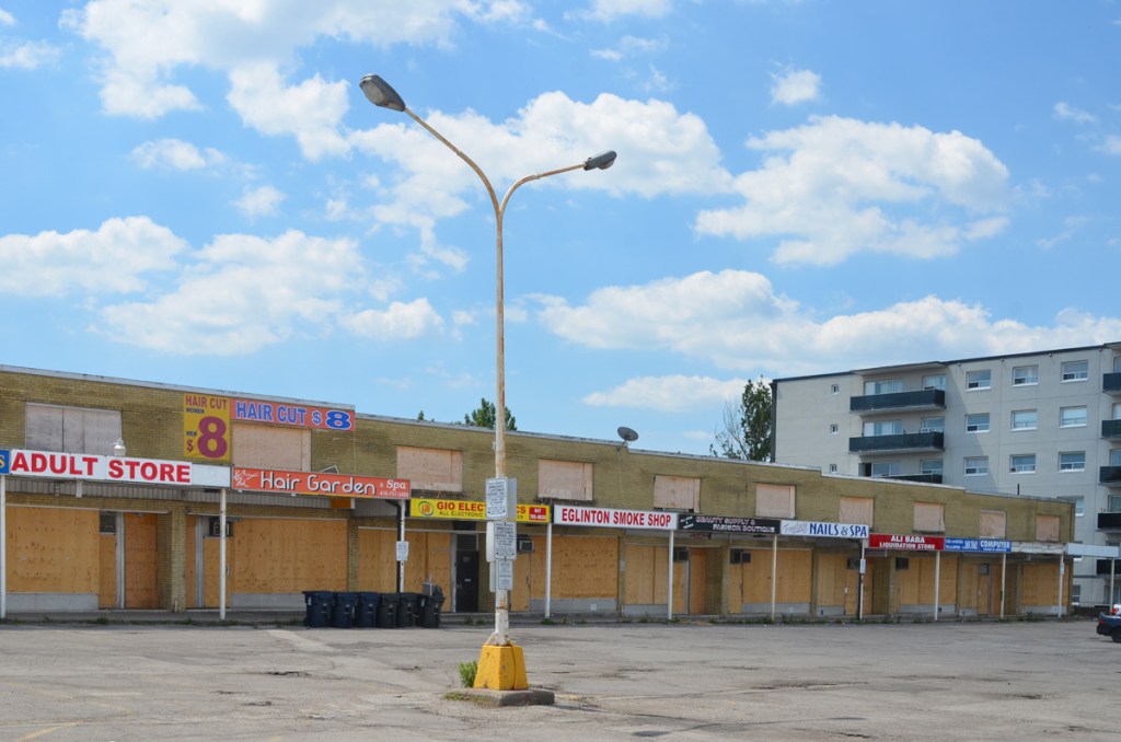 old overhead light standard in an empty parking lot in front of a boarded up strip mall with Adult store, hair garden, smoke shop, nails spa and ali baba restaurant
