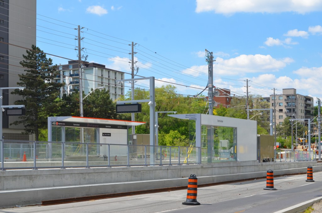 New Ionview LRT station with its  protected areas, like a bus shelter, at surface level, between the lanes of traffic on Eglinton, apartment buildings in the background