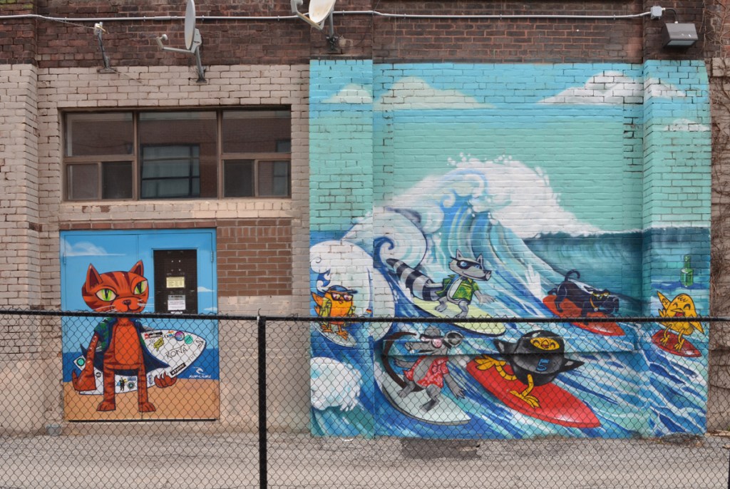 A mural by Uber 5000 in 2 parts.  on the left is a tabby cat holding a surf board.  on the right are some characters surfing, 3 yellow birdies, on in a wet suit, and three dogs. 