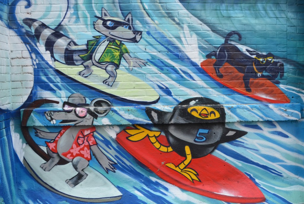 surfing mural by Uber 5000, yellow birdie in a black wet suit, a dog in a green Hawaiian shirt, a dog with goggles