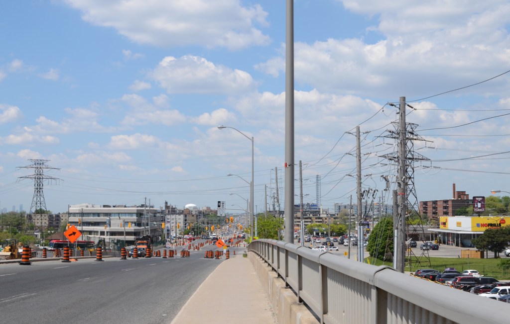 view along Eglinton westward towards Kennedy Road, wide street, hydro poles, low rise development, lots of black and orange construction cones