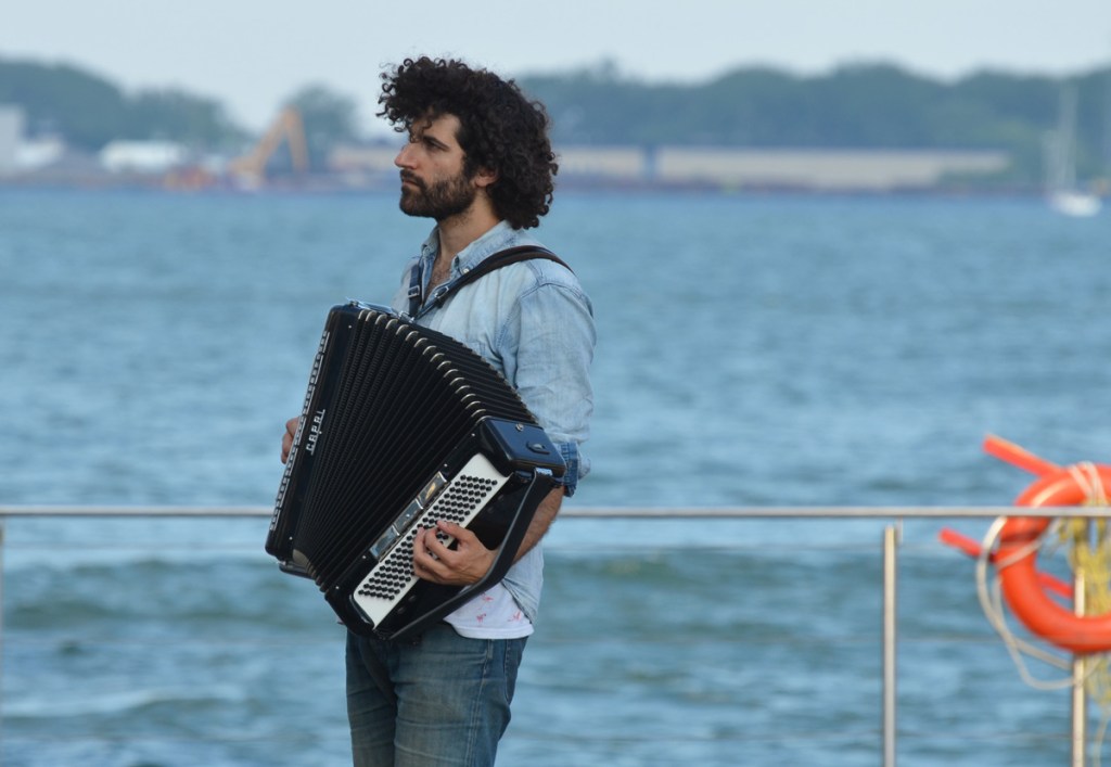 A male accordion player, Joseph Landau, stands beside the waterfront. Curly black hair.