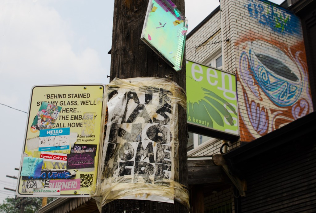 signs on a wood utility pole in front of a painting on a brick wall of a cup of coffee. A poster with words Hate has no home here. An ad for a store that is partially obscured by the pole, and a fake street sign that starts with Behind Stained steamy glass, we'll... and the rest is blocked by many stickers