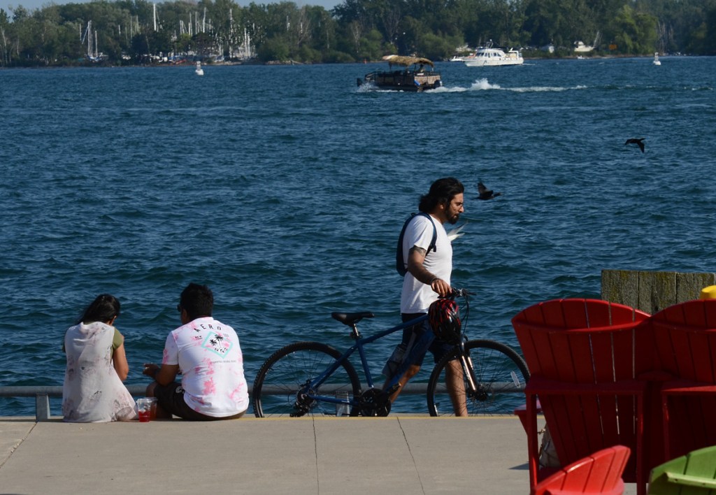 A couple sit with their backs to the camera on the edge of Lake Ontario while a male cyclist walks his bike past. Two red Muskoka chairs in the picture as well as boats on the water