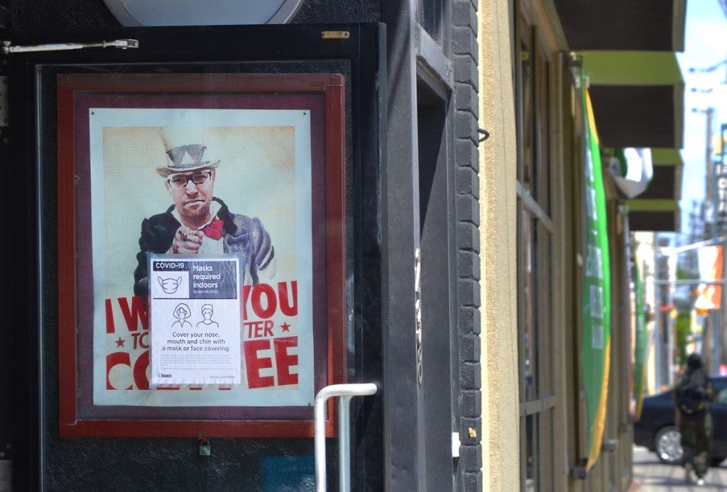 poster of a man, parody of uncle sam poster, top hat and pointing, words say I want you to like better coffee. On the exterior of a door to a coffee shop, with covid mask notice in front of it