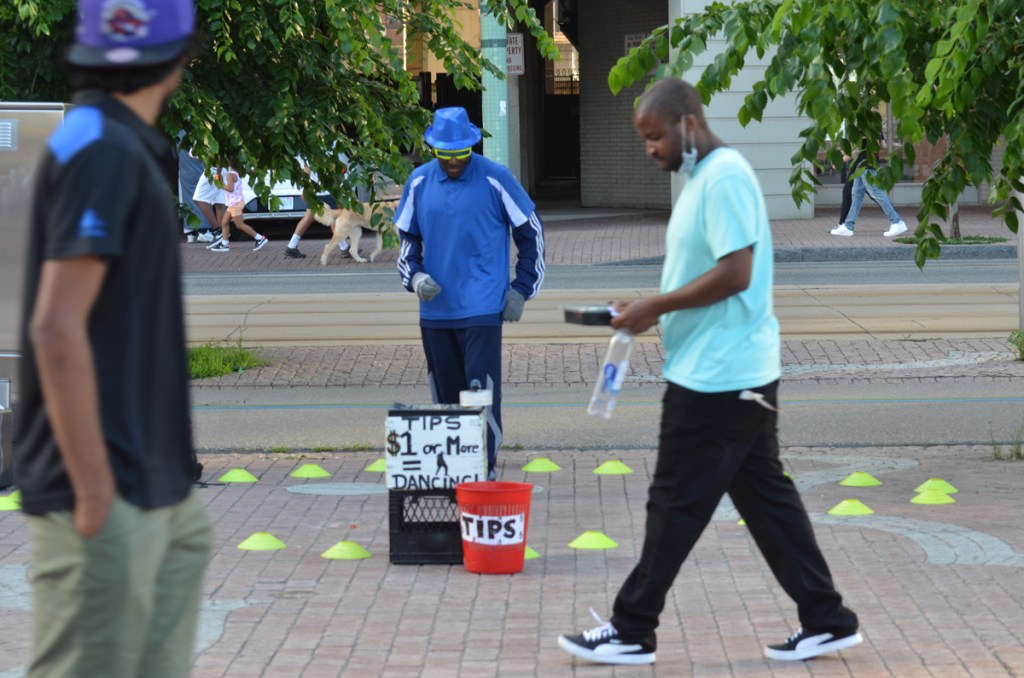 Two men walk past another man dressed in blue who dances for tips. He's standing still because no one has given him any money