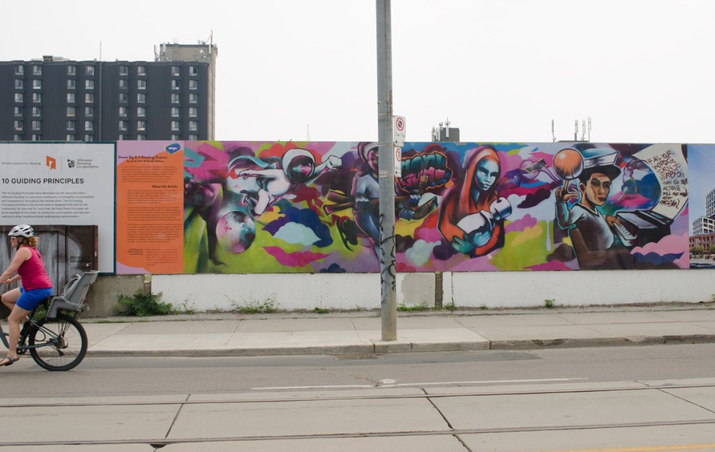 a woman on a bicycle rides past hoardings around Alexandria Park redevelopment, with a mural on it by Javid Jah and Danile Deluxe