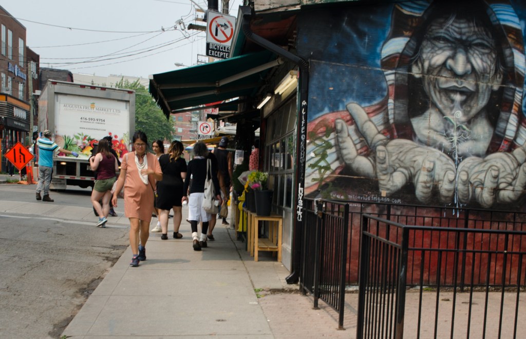a woman in a coral coloured dress walks past a mural of an old woman with her hands outstretched, asking for something, a red and white shawl over her head