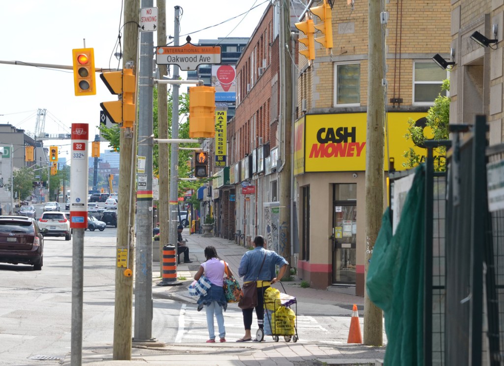 a mother and daughter stand with a shopping buggy waiting for a traffic light on Eglinton West at Oakwood, stores, traffic, man on bench on sidewalk