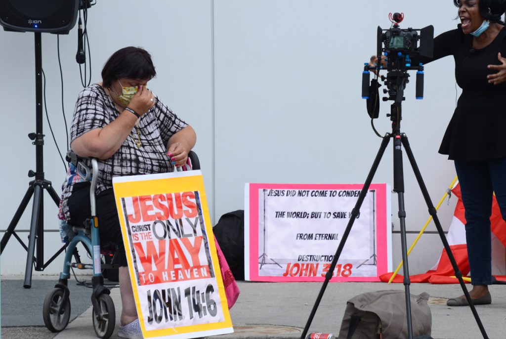 a black woman being video'd, a woman in a wheelchair with a sign leaning against her knees that says Jesus Christ is the only way to heaven John 14: 6 Sign at the back says Jesus did not come to condemn the world but to save it from eternal destruction.
