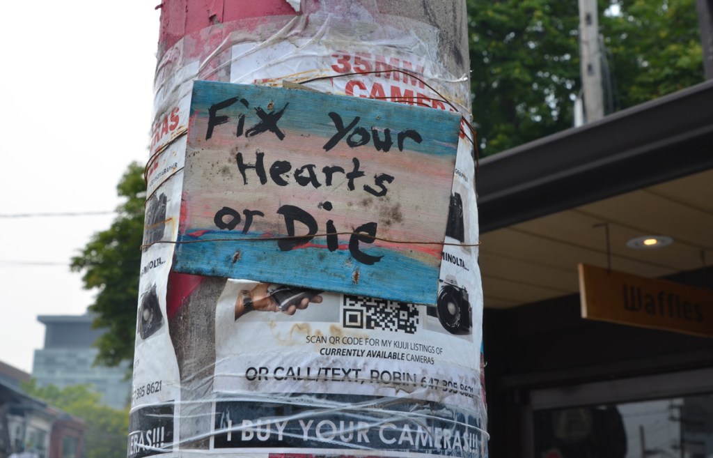 graffiti sign on a pole, covering an add for 35 mm cameras for sale, words that say fix your hearts or die