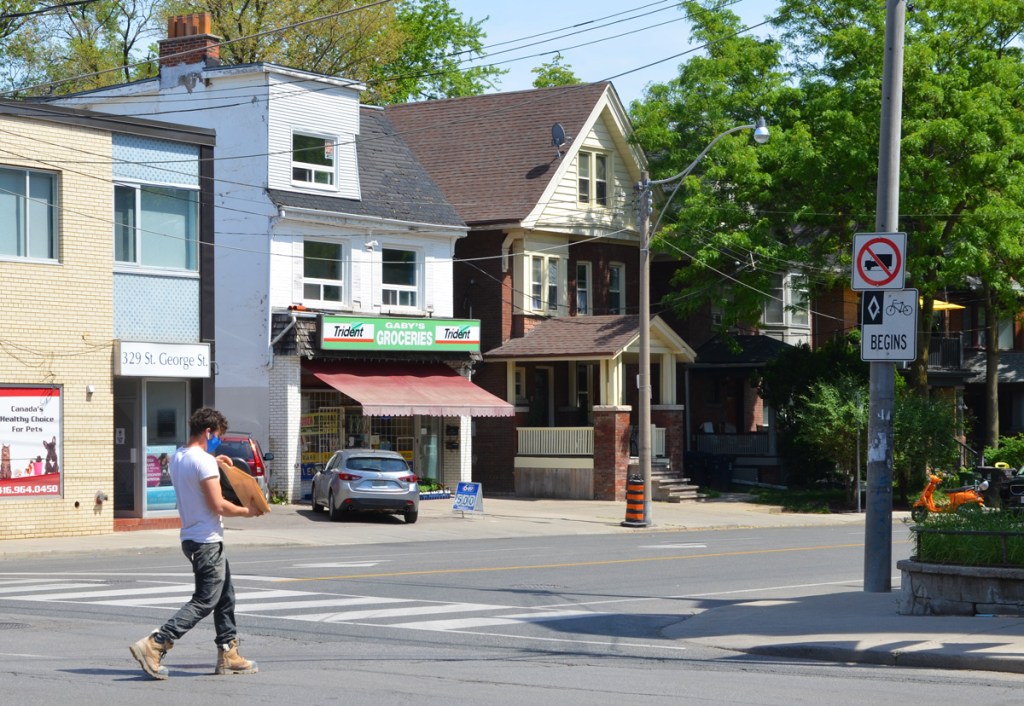 a man crosses Dupont near the intersection with St. George Street, Gabys Groceries is in the background with its green Trident gum advertisement sign