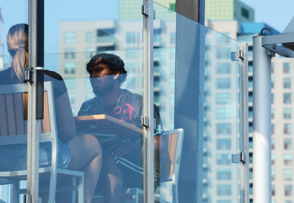 A couple sits at a table on an upper level balcony patio, behind glass barrier. With condo buildings behind