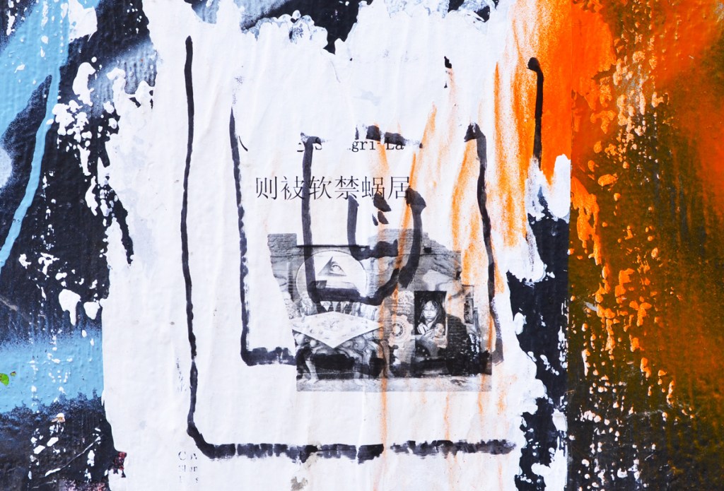 paper paste up graffiti on a concrete pole with chinese lettering and black and white photos on it, orange and blue spray paint beside the paste up 