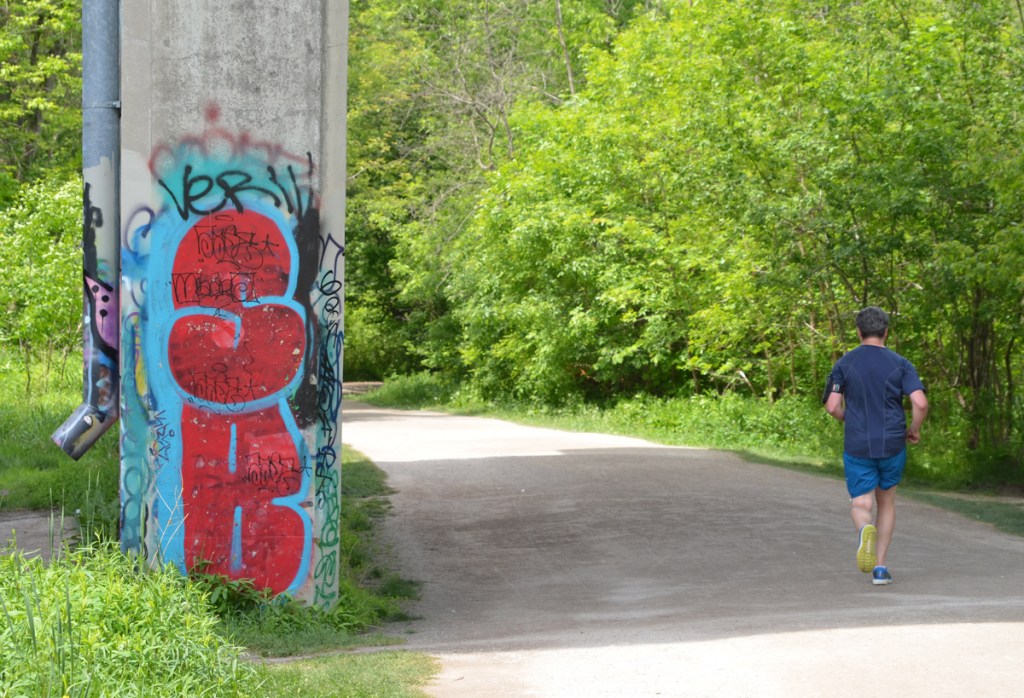 a man in blue top and shorts runs on a path under a bridge ,  support of bridge has graffiti on it, large red s and b on blue background 