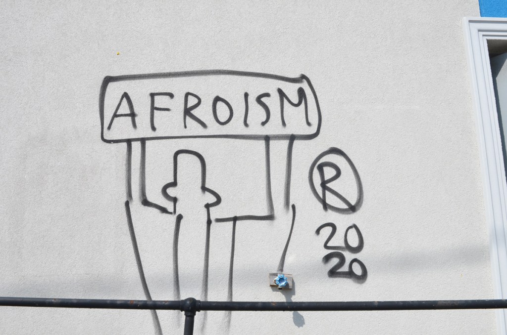 graffiti, black spray paint on a light grey concrete wall, outline of a person holding up a sign above head that says Afroism, 2020
