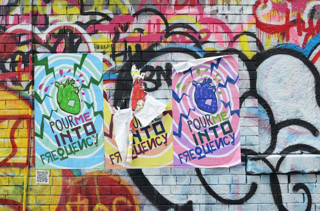 3 large graffiti posters in an alley, covering other street art, each with an anatomically correct heart in different colours (red, green and blue), and the words pour me into frequency