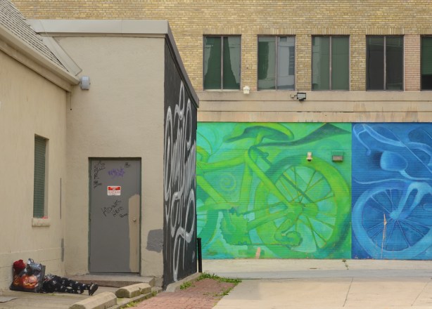 alley behind Ryerson Image Centre with large murals of green bike and blue bike, also with a person asleep in the alley by the church 