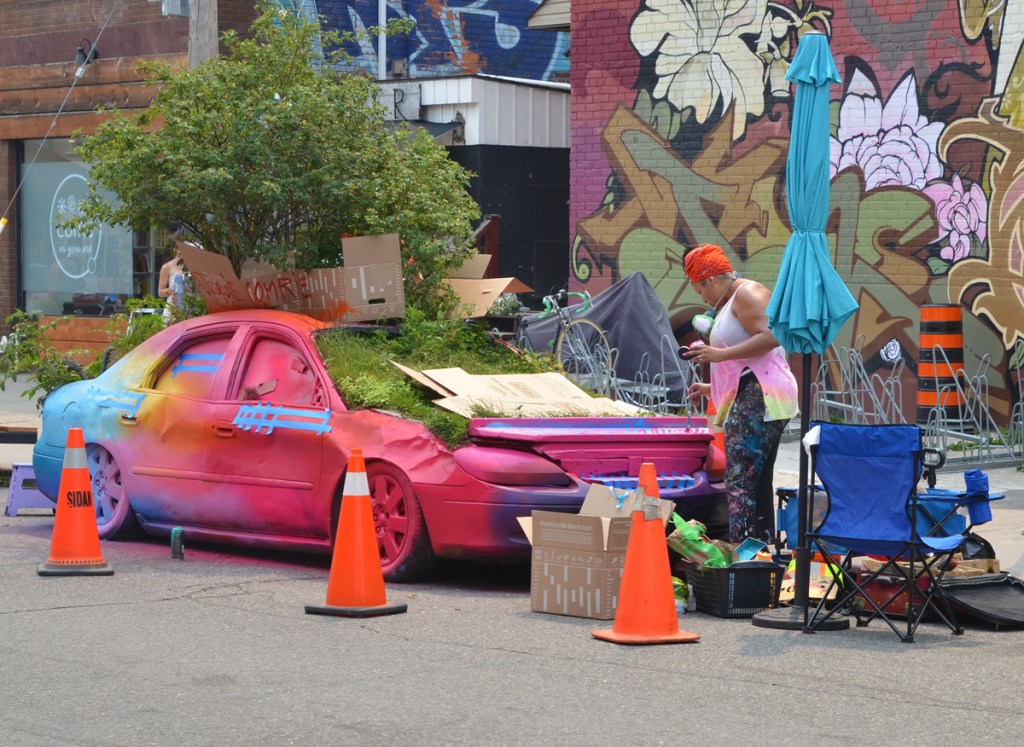 woman wearing an orange turban-like hat is painting a car in many bright colours, the car is full of plants growing inside it