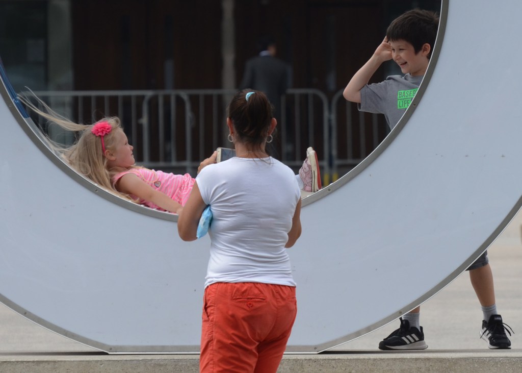 a young boy teases his sister as she is lying in the center of the O of the Toronto 3 d sign, mother is taking a picture of daughter. mother's back is to the camera