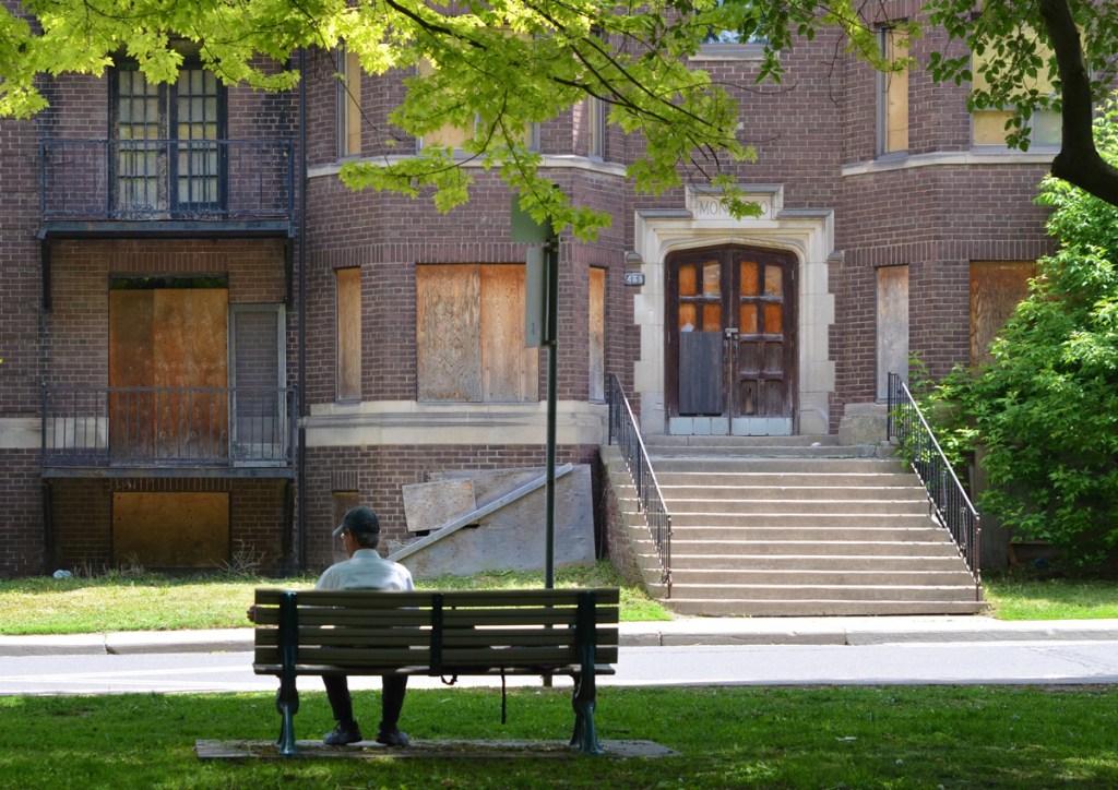 man sitting on a bench in a park with back to camera, looking across street to old boarded up brick apartment building