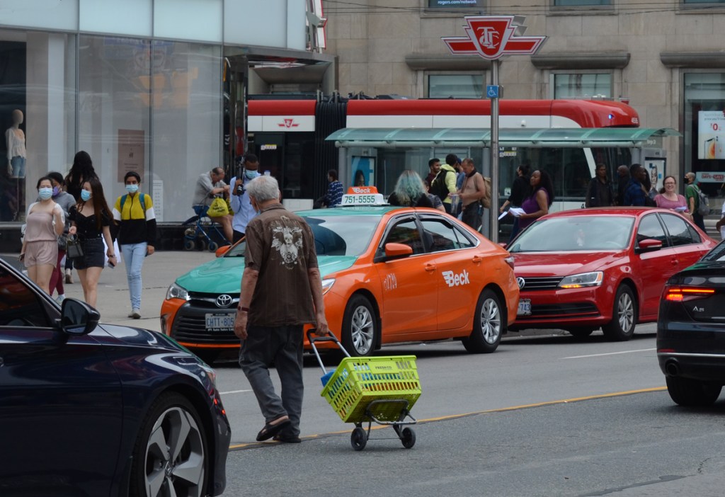 an older man in a brown shirt crosses Yonge street traffic while pulling a green basket on a dolly. people in masks walking on the sidewalk, a Beck taxi parked on Yonge, a bare chested man by the bus stop, other people
