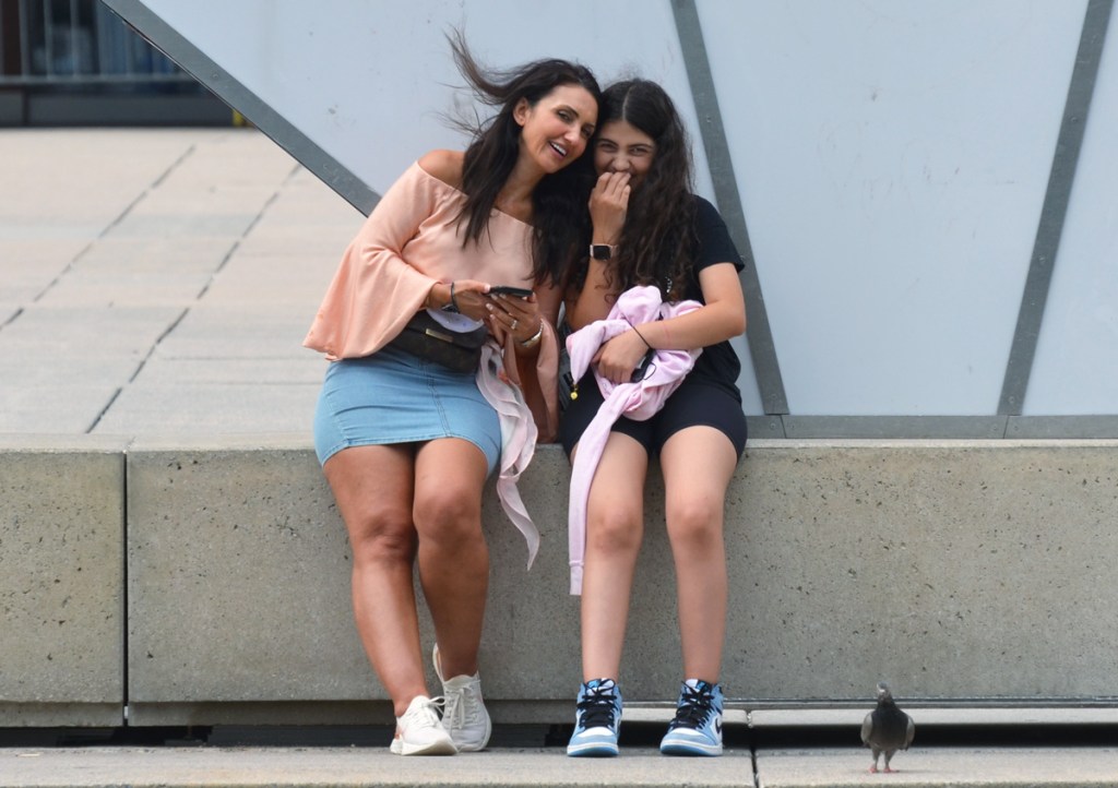 a mother and daughter, both with long black hair, sit in front of the toronto 3 d sign, leaning in together, smiling, a pigeon stands by their feet