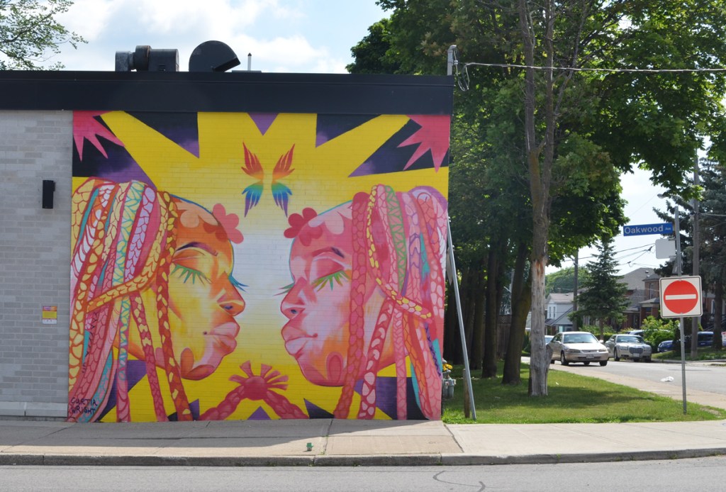 mural on the side of The Beer Store, in bright colours, two women with dreadlocks, eyes closed and about to kiss 
