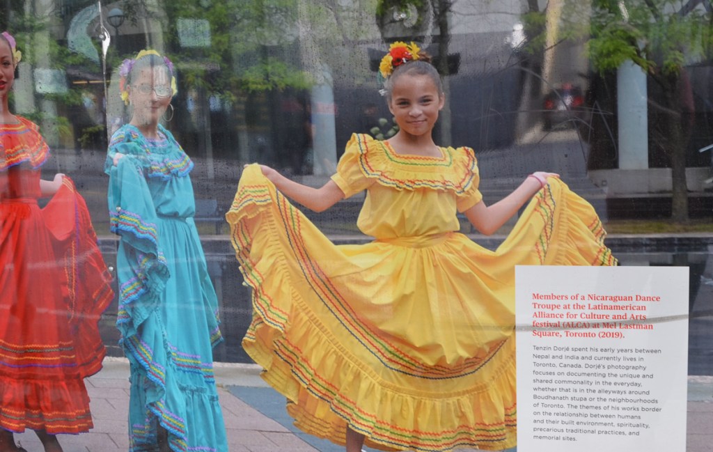 photograph on hoardings, three women of Nicaraguan dance troupe in long colourful dresses, red, yellow, and blue, also with flowers in their hair 