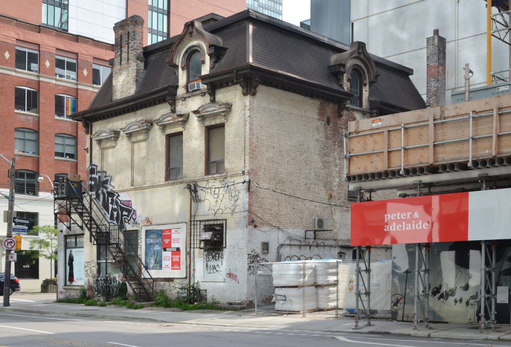 old pale beige brick house from the late 1850s with brown mansard roof at the northwest  corner of Peter and Adelaide.  empty, boarded up, with some tags on it, metal exterior staircase from ground level to second story, Second Empire style