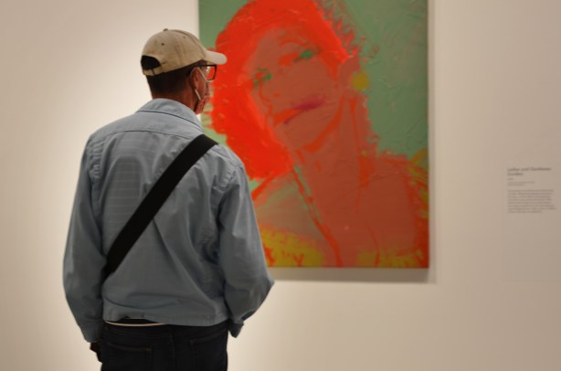a man in pale blue jacket and baseball cap stands in front of a portrait by Andy Warhol 